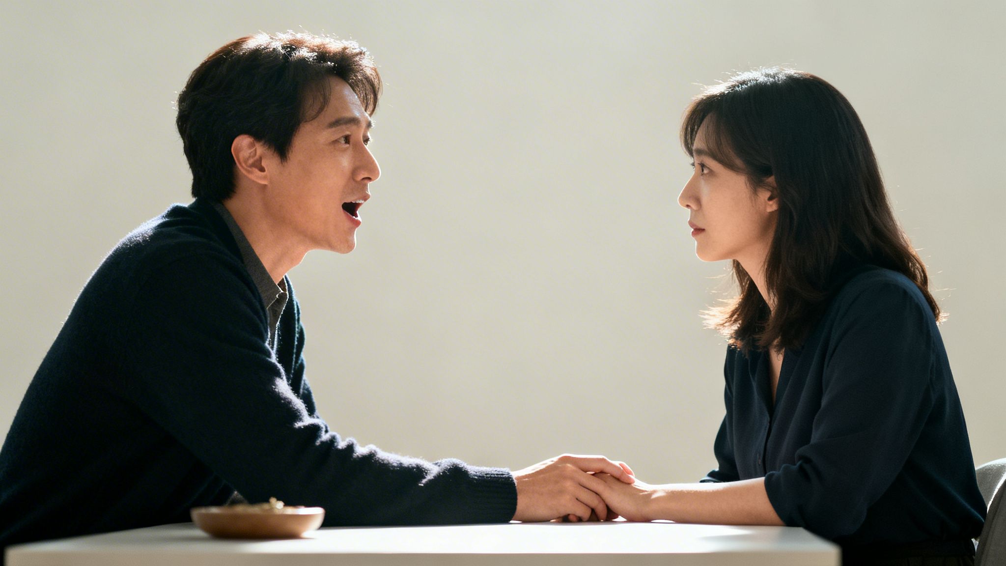 Profile view of a man speaking to a woman, holding hands across a table.