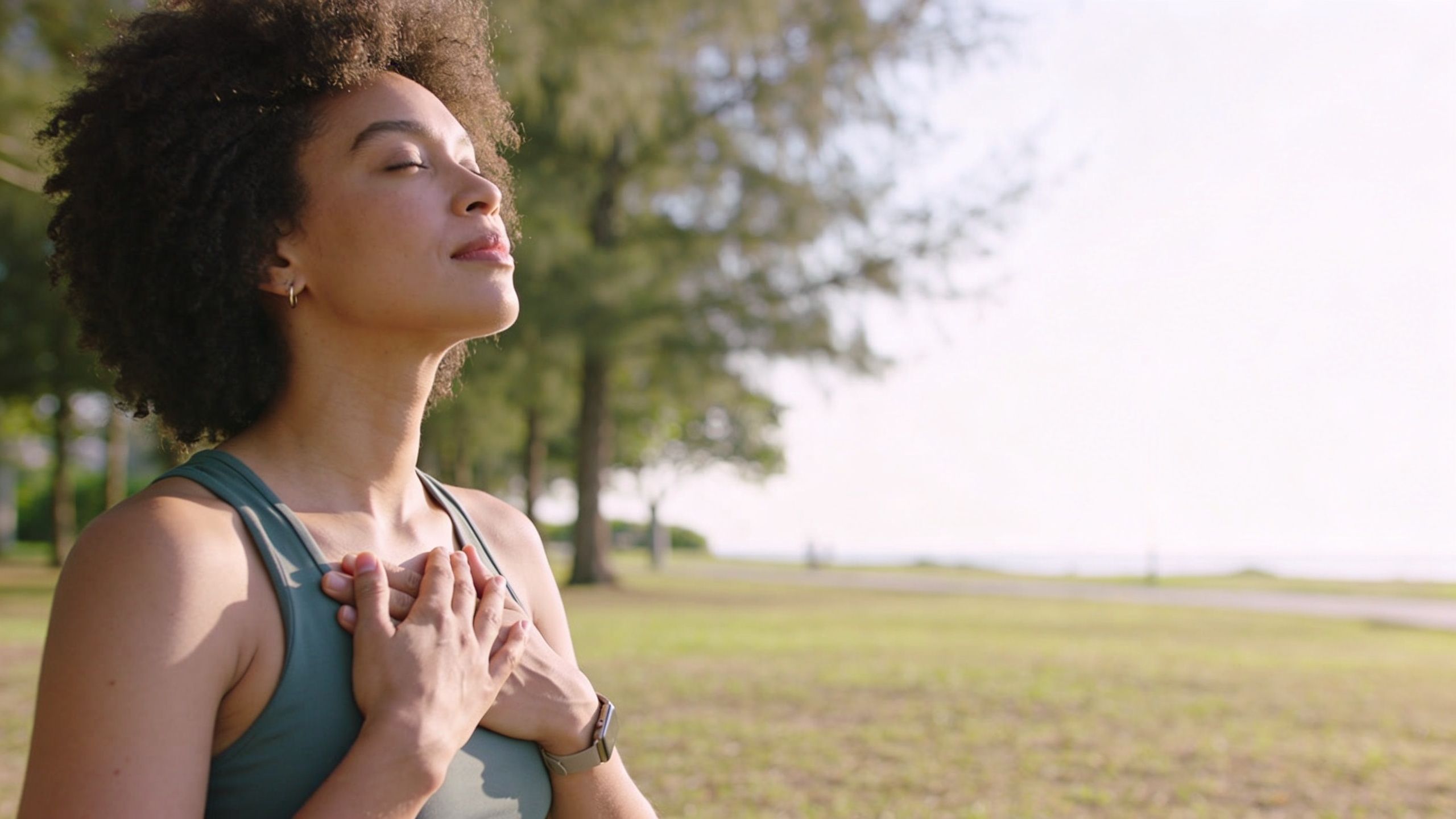 A woman with curly hair practicing mindfulness and breathing exercises outdoors in a park during the day.