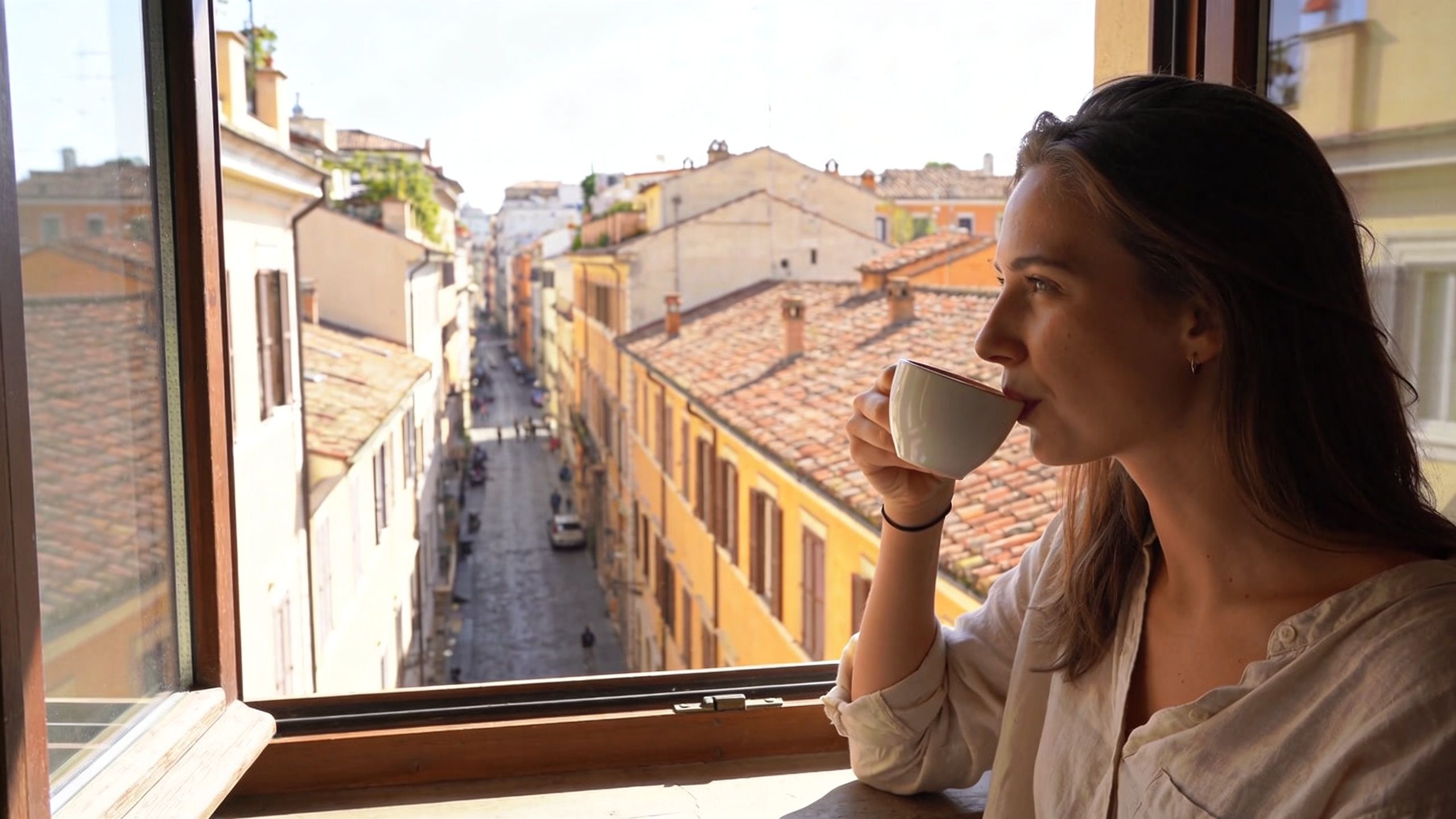A woman looks out a window at a street in Rome while drinking coffee, embodying an expat lifestyle.