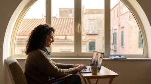 A young international woman in her late 20s sits near a large window with soft Naples afternoon light, having a calm online therapy