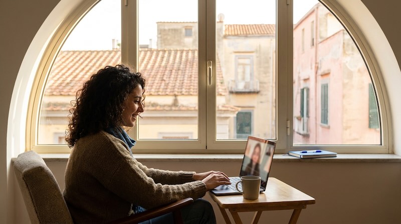 A young international woman in her late 20s sits near a large window with soft Naples afternoon light, having a calm online therapy
