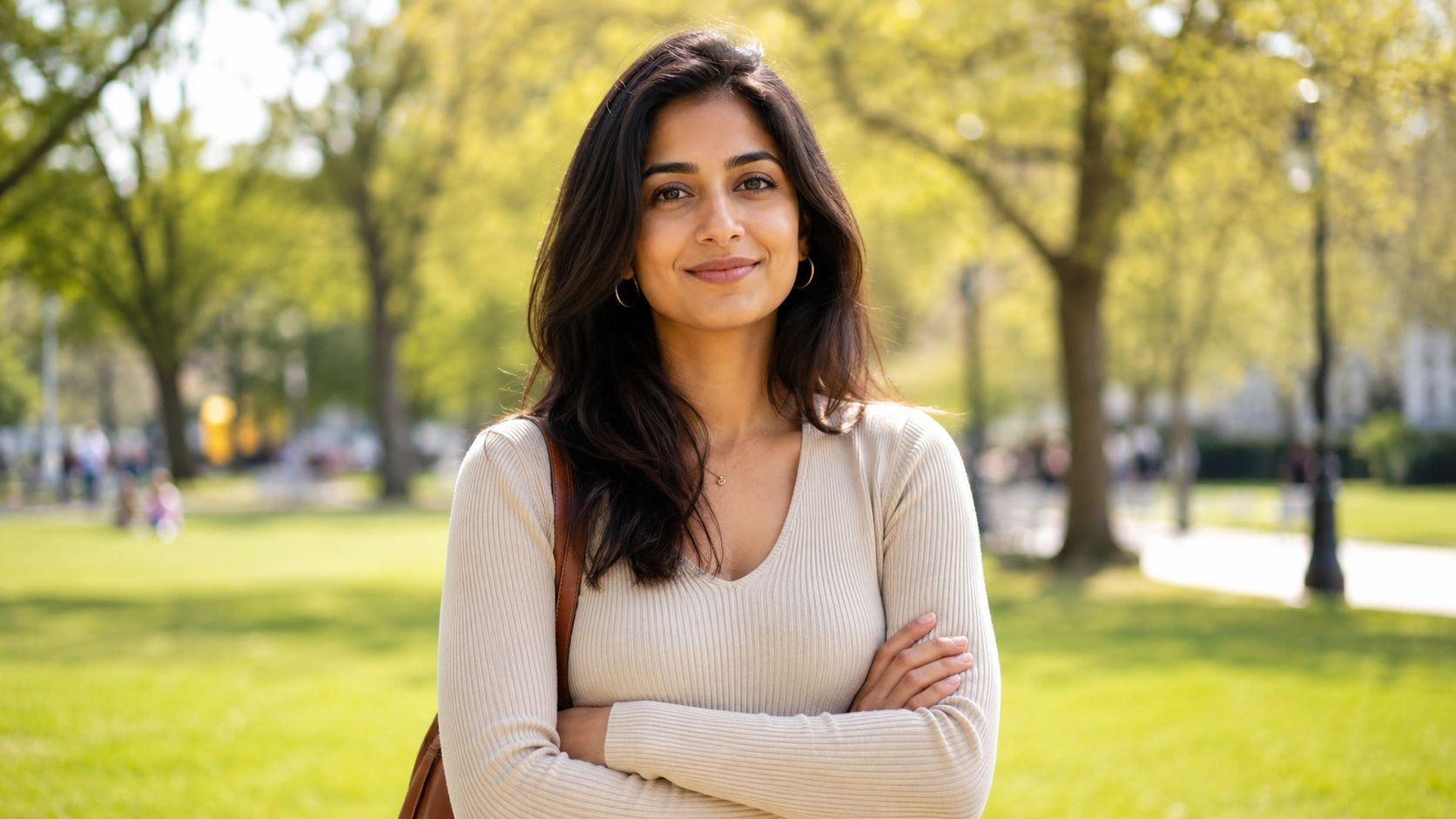 A smiling young woman with long dark hair standing in a park with arms crossed.