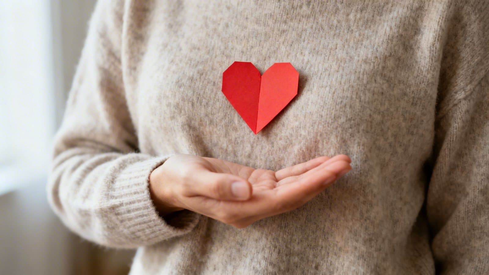 A person in a beige sweater with a red origami heart on their chest, offering an open hand.